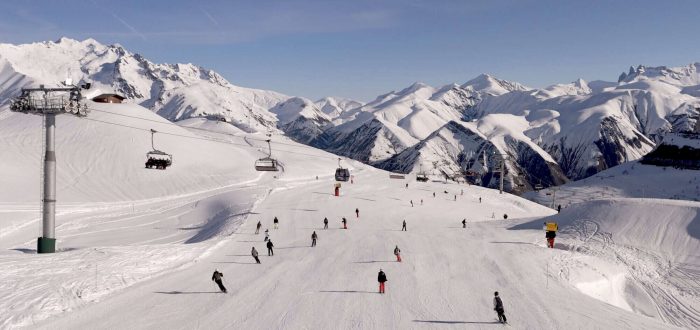An aerial view showing skiers on a piste, with a ski lift and impressive mountain range in the background.