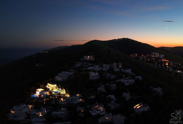 Coastal village illuminated at dusk with hillside homes and ocean view.