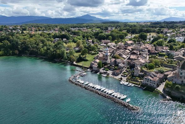 Aerial view of Yvoire on the coast of Lake Geneva, with boats docked, lush greenery and a scenic mountain backdrop.